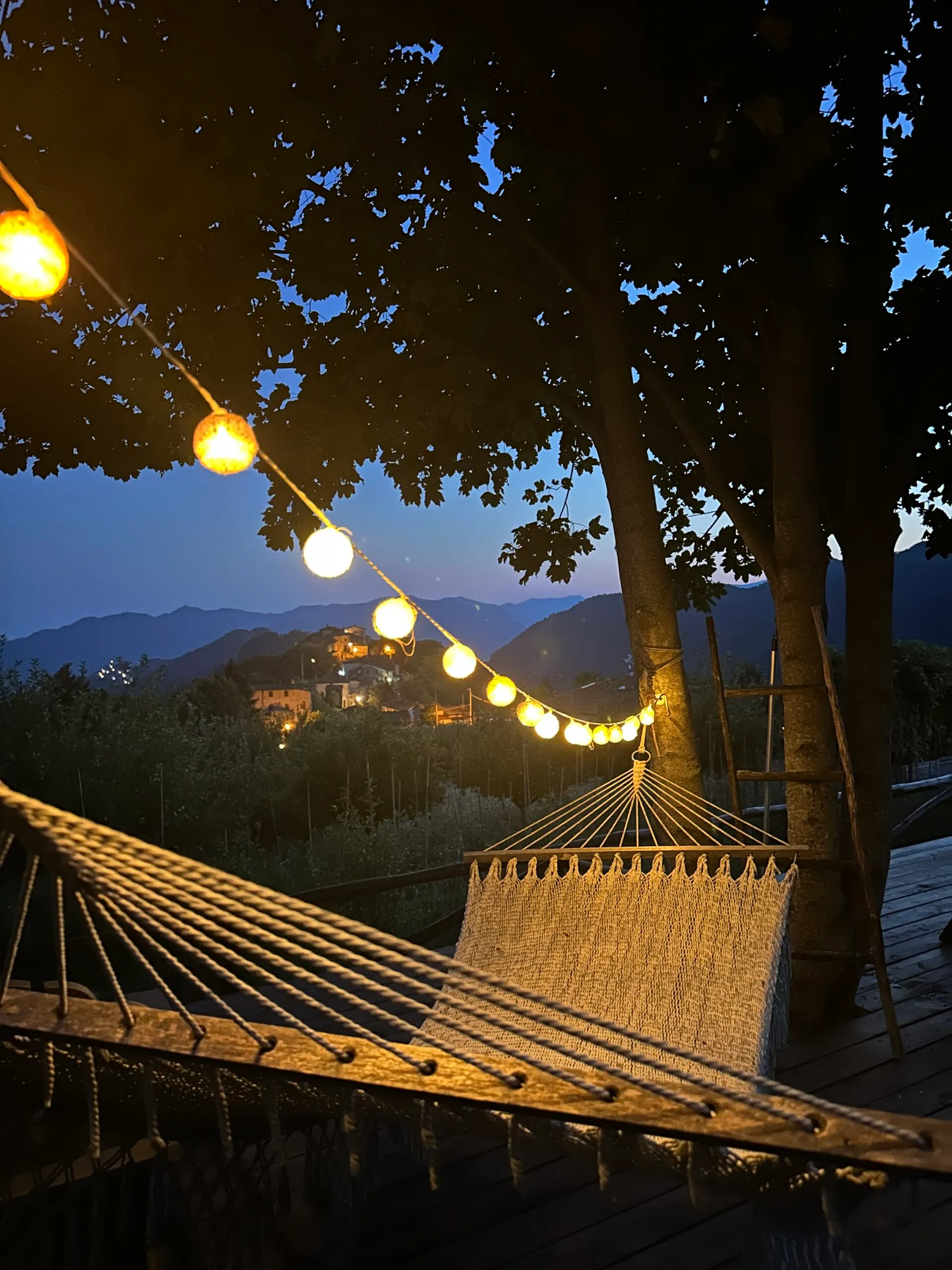 Hangmat op het terras bij schemer, met lichtjes in de boom en het verlichte dorp op de heuvel