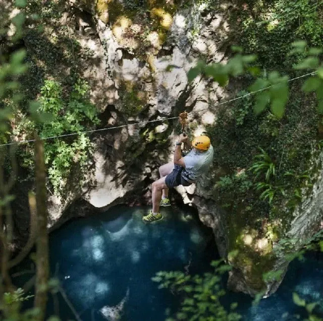 Iemand met helm aan een kabel boven een turkooizen poel in een kloof